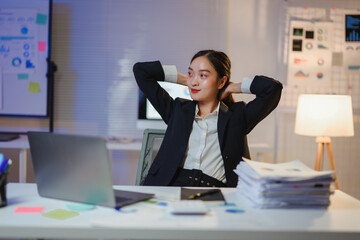 Female professional stretching arms near laptop, relaxing during late-night work session in dimly...