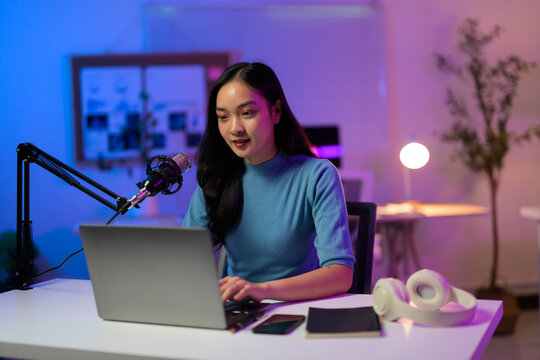 Asian female podcaster speaking into professional microphone while working remotely from home office, laptop nearby
