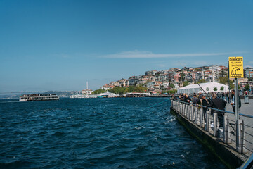 Naklejka premium Scenic waterfront promenade with people, boats, and a yellow Turkish warning sign by the blue sea under clear sky. Vibrant urban life, leisure, travel, and safety.