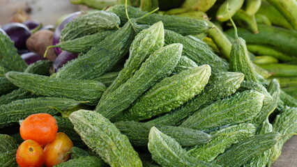 Vibrant Farmers Market Display Featuring a Large Pile of Fresh, Bumpy Green Bitter Gourds alongside Tomatoes and Eggplants
