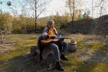 Blonde woman playing string guitar outdoors in autumn forest. Concept of sound therapy, mental...
