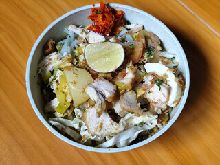 A bowl of Indonesian chicken soto with shredded chicken, sliced boiled egg, vermicelli, sliced cabbage, bean sprouts, celery, chili sauce and lime, mixed with delicious sauce. On a wooden table.	
