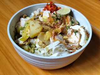 A bowl of Indonesian chicken soto with shredded chicken, sliced boiled egg, vermicelli, sliced cabbage, bean sprouts, celery, chili sauce and lime, mixed with delicious sauce. On a wooden table.	
