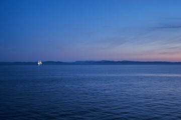 A distant ferry illuminated on the calm waters of Vancouver at twilight with mountains in the...