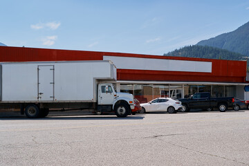 A large white delivery truck and several cars parked outside a red commercial building with...
