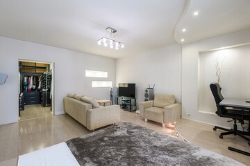 bright, modern living room features a couch, armchair, TV setup, and a desk area. A walk-in closet is visible. The decor is minimalist and clean