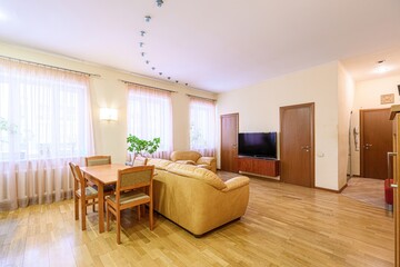 cozy living room with a wooden floor, a dining table with four chairs, a leather sofa, a TV, and three windows with sheer curtains