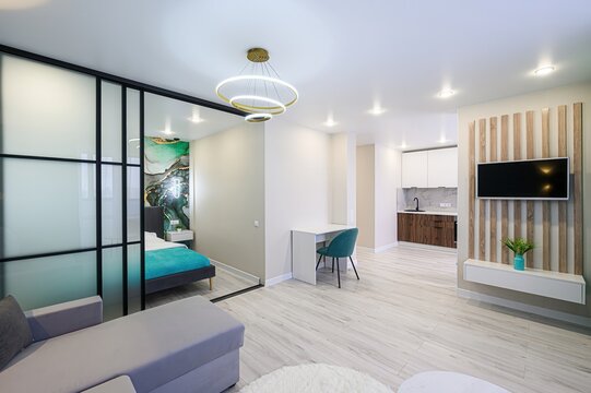 Modern apartment interior with bedroom separated by glass partition, kitchen area, TV wall, and contemporary furniture