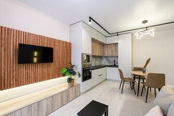 Modern apartment with TV on wood-paneled wall, sleek kitchen cabinets, and a cozy dining area....