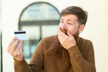 Redhead man with beard holding a credit card at outdoors with surprise and shocked facial expression