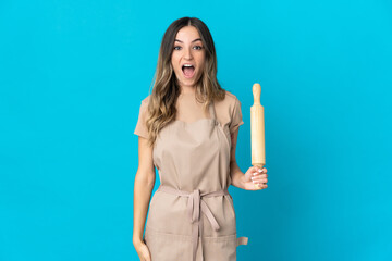 Young Romanian woman holding a rolling pin isolated on blue background with surprise facial...