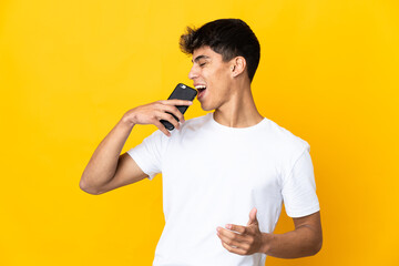 Young man over isolated yellow background using mobile phone and singing
