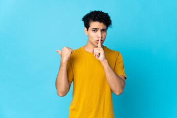 Young Venezuelan man isolated on blue background pointing to the side and doing silence gesture