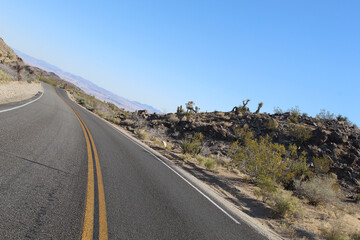 Scenic desert highway with yellow center line curving through arid rocky landscape near Joshua Tree National Park, California, USA. Clear blue sky and wilderness in view.