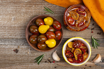 Flat lay photo of a glass jar and and saucer with sun dried red and yellow tomatoes in olive oil and with herbs, wooden bowl with cherry tomatoes, rosemary, garlic, pepper in the wooden table.