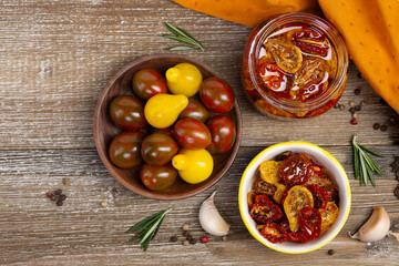 Flat lay photo of a glass jar and and saucer with sun dried red and yellow tomatoes in olive oil and with herbs, wooden bowl with cherry tomatoes, rosemary, garlic, pepper in the wooden table.