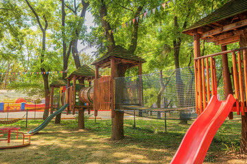 A colorful wooden playground with slides, climbing nets, and tunnels set among trees in a sunny park decorated with festive banners.