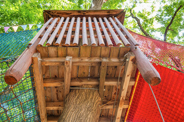A wooden treehouse with colorful rope bridges and safety nets surrounded by lush green trees in a sunny outdoor playground.