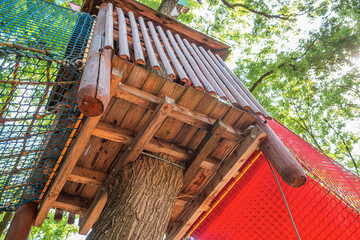A wooden treehouse with colorful rope bridges and safety nets surrounded by lush green trees in a sunny outdoor playground.