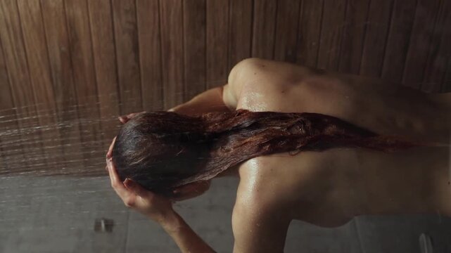 Drops of water fall on a young naked girl in the shower in slow motion. A young woman takes a shower. Concept of daily self-care. Body care concept. Beauty routine. Close-up. Vertical footage.