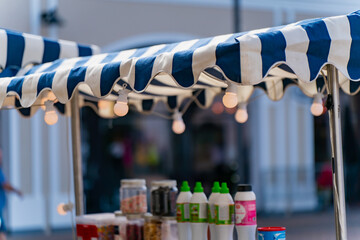 Outdoor drink stand with striped awning and glowing bulbs, festival market stall closeup showing refreshment bottles under canopy, cozy summer evening street fair beverage kiosk