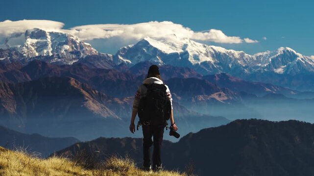 A man walks in front of dramatic mountain views on the Himalayas, Nepal. He is wearing a backpack, a woollen hat and is holding a camera. He brings the camera to his face to take photos.