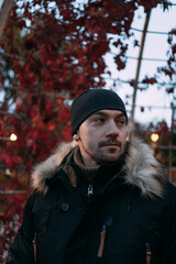 Portrait of a man in black winter coat with fur hood and patterned gloves outdoors in autumn park....