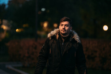 Obraz premium Person in black winter coat with fur hood standing outdoors in evening park. Warm clothing, cozy autumn atmosphere, street lights, modern fashion, and urban night stroll