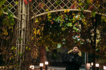 Man in black winter coat with fur hood and gloves standing under autumn arch in park at night,...