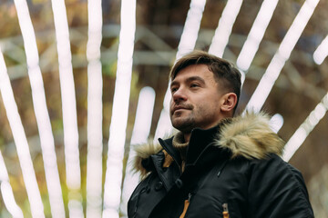 Man in a black winter coat with fur hood and sweater standing outdoors under bright decorative...