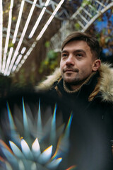 Close-up of a man in a black winter jacket with a fur hood against a background of geometric light patterns. Urban night, creative atmosphere, modern street style, autumn mood