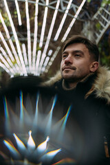 Close-up of a man in a black winter jacket with a fur hood against a background of geometric light patterns. Urban night, creative atmosphere, modern street style, autumn mood