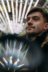 Close-up of a man in a black winter jacket with a fur hood against a background of geometric light patterns. Urban night, creative atmosphere, modern street style, autumn mood