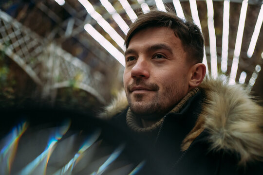 Fototapeta Close-up of a man in a black winter jacket with a fur hood against a background of geometric light patterns. Urban night, creative atmosphere, modern street style, autumn mood