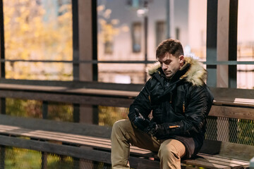 A man in a black winter jacket with a fur hood, beige pants, and boots sits casually on a bench outdoors at night. Urban background, autumn atmosphere, modern street style