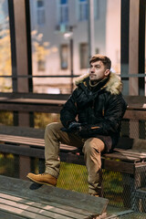 A man in a black winter jacket with a fur hood, beige pants, and boots sits casually on a bench outdoors at night. Urban background, autumn atmosphere, modern street style