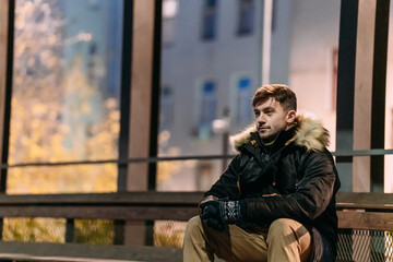 A man in a black winter jacket with a fur hood, beige pants, and boots sits casually on a bench outdoors at night. Urban background, autumn atmosphere, modern street style