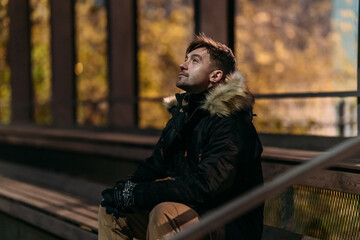 A man in a black winter jacket with a fur hood sits on a bench under a canopy in a park at night....