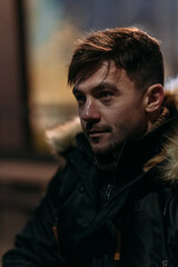 A man in a black winter jacket with a fur hood is outdoors at night. Warm autumn tones and an urban background highlight modern casual fashion and a cozy city evening atmosphere