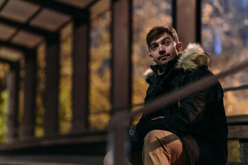 A man in a black winter jacket with a fur hood is outdoors at night. Warm autumn tones and an urban background highlight modern casual fashion and a cozy city evening atmosphere