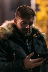 A man in a black winter jacket with a fur hood holds a smartphone outdoors at night. Warm autumn colors and urban background create a cozy, stylish evening city mood
