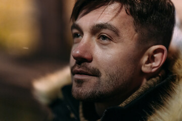 Close-up of a man in a black winter jacket with a fur hood outdoors at night. Warm autumn lighting and cozy atmosphere highlight urban fashion and seasonal style