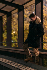 A man in a black winter jacket with a fur hood, beige pants, and boots stands on a wooden bench under a canopy in a park at night. Cozy autumn mood, modern casual style