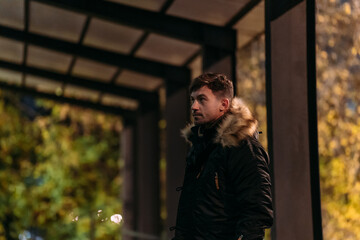 A man in a black winter jacket with a fur hood stands under a canopy in a park at night. Warm lights, autumn foliage, and modern style create a cozy, fashionable evening scene