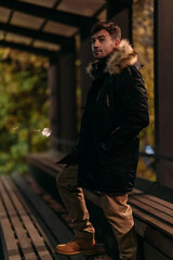 A man in a black winter jacket with a fur hood stands stylishly on a wooden bench under a canopy in a park at night. Autumn mood, cozy atmosphere, casual pose, warm clothing