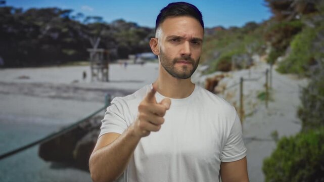 Young hispanic man pointing at camera with a serious expression on a beach, surrounded by sea and nature, wearing a casual white shirt outdoors.