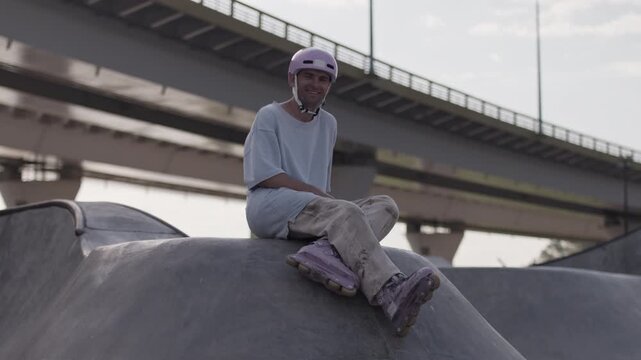 Outgoing inline skater smiles while sitting on ramp top by bridge