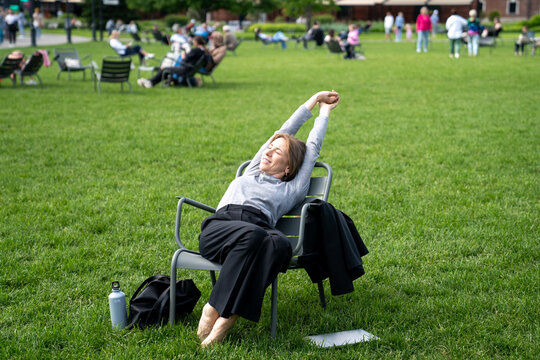 Laid-back lazy and drowsy woman stretching and napping in park lounger during hot summer day. Siesta, idleness and inability to work in warm weather and relaxation in green park with trees and grass