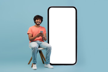 Cheerful curly young eastern guy in casual outfit enjoying newest mobile application, sitting on...