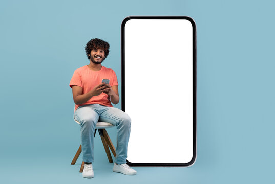 Cheerful curly young eastern guy in casual outfit enjoying newest mobile application, sitting on chair by big smartphone with white empty screen, using cell phone and smiling, mockup, copy space